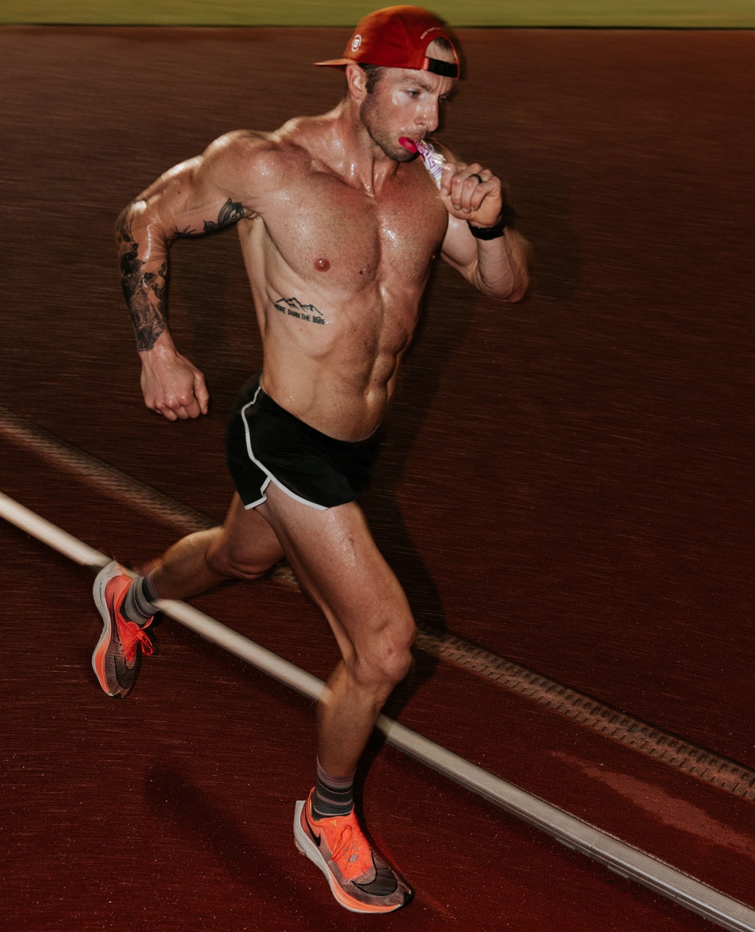 Man running on a track at night, wearing a red cap and holding a snack.