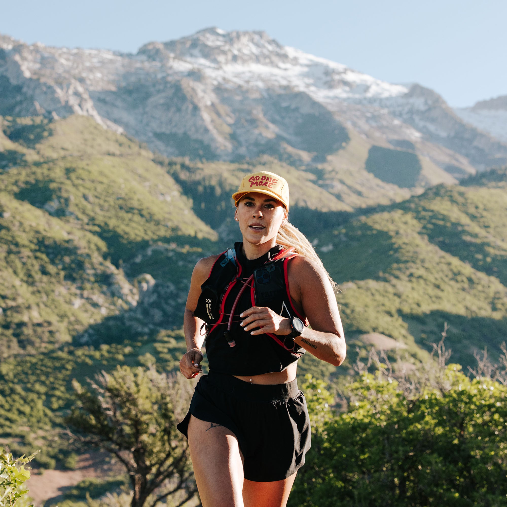 A woman running on a mountainous trail with green hills and snow-capped peaks in the background.