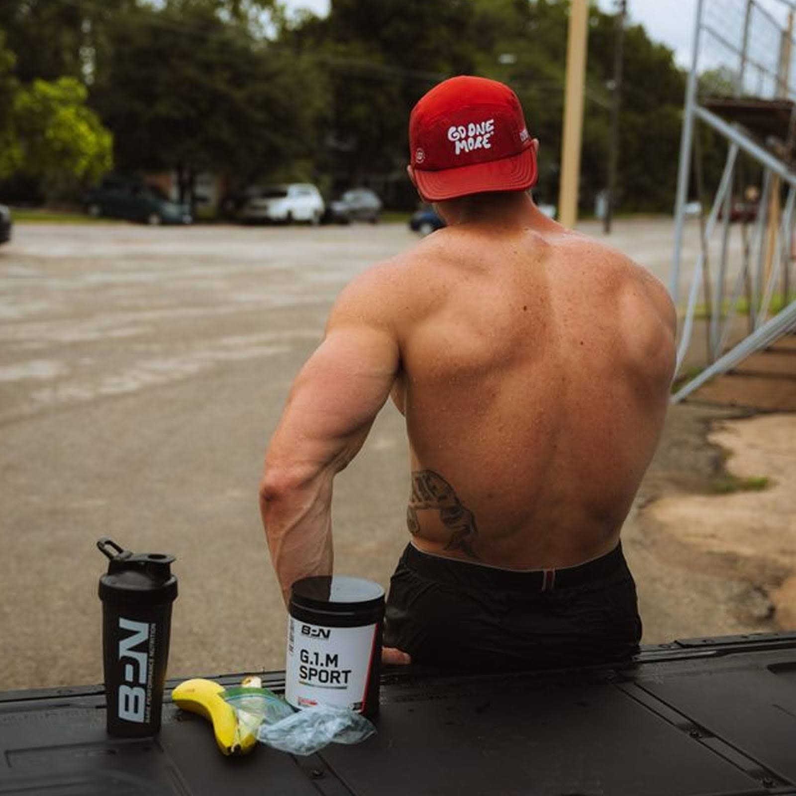 Shirtless man with cap sits outdoors, gym supplements and a banana are on the table in front of him.
