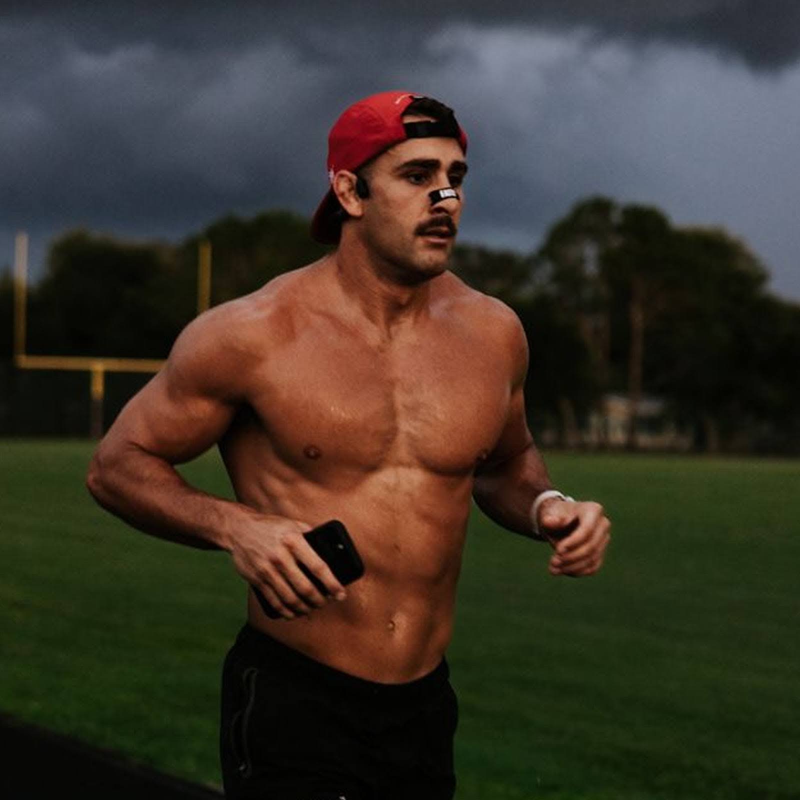Shirtless man running on a field, wearing a red cap and nasal strip.