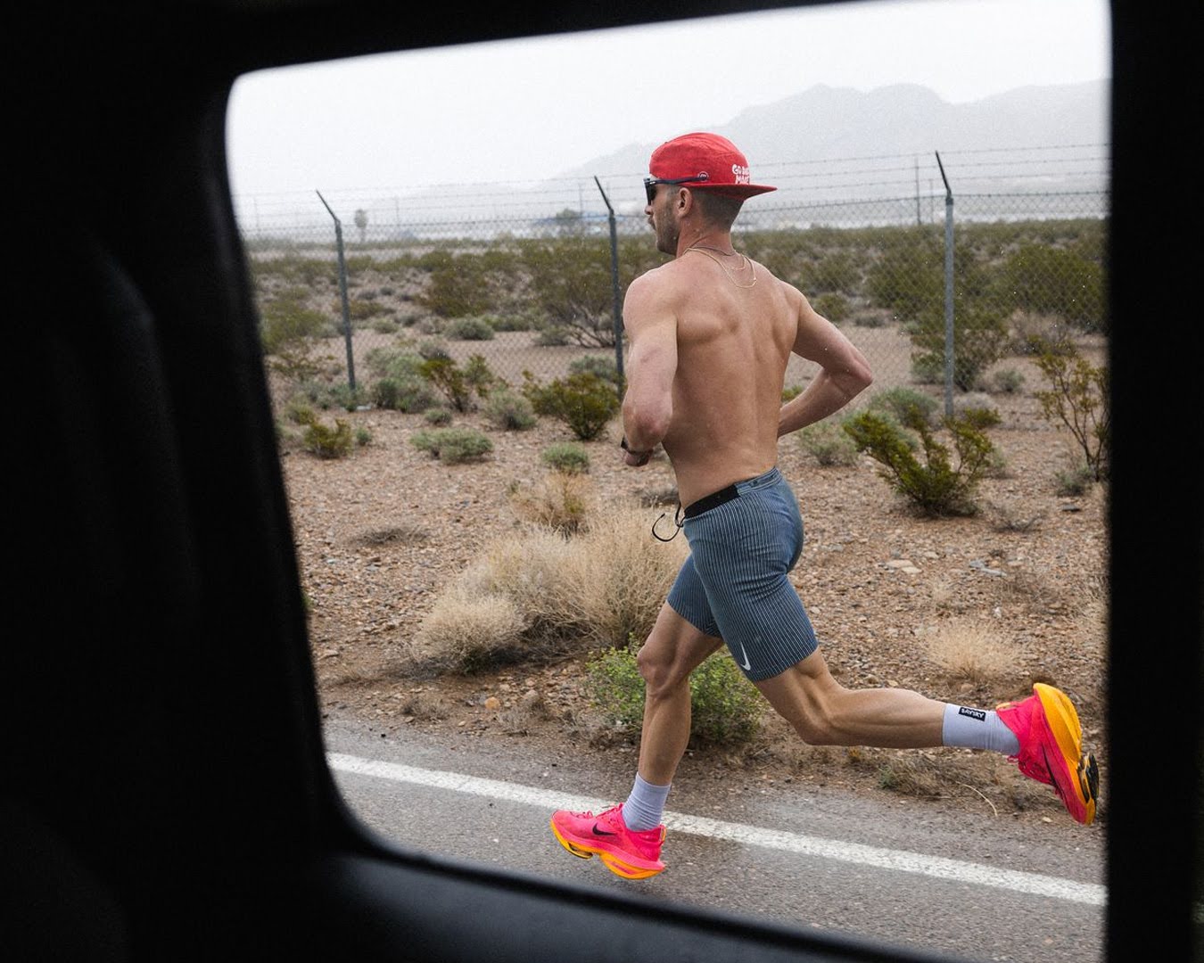 Shirtless man with red cap and pink shoes running on roadside near fence.